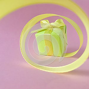 Green gift box at the end of the spiral yellow ribbon,  pink background, square.