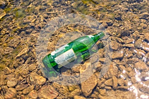 Green glass bottle lying in the shallow water of the river. Concept of environmental pollution. Close up