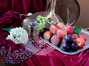 Green grapes, plums, peaches, nectarines, jugs, a glass of wine, white hydrangea, a Dutch still life on a silk tablecloth, with
