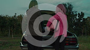 Green hair girl and her girlfriend with pink hair opening the car's trunk outdoors