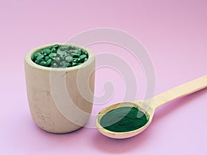 Green hawaiian spirulina in tablespoons pills and powder on light lilac background.