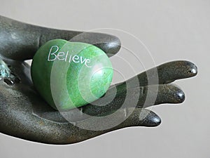 Green Heart Shaped Rock with the word BELIEVE etched in it held in a decorative hand statue