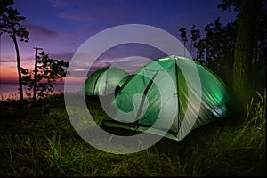 Green illuminated from inside tent above river at sunset.