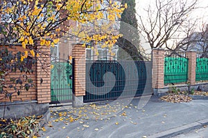 Green iron gate with forged pattern and part of the fence made of metal and brown bricks on the street