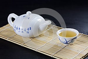 Green japanese tea on a bamboo mat on black stone, one white cup of tea and teapot with green tea view from above