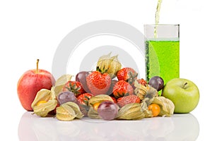 Green juice pouring in to the glass behide red and green apples with orange physalis, purple grapes and red strawberries on white