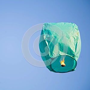 Green lampion rising on the clear blue sky with a flame lit underneath it - Square