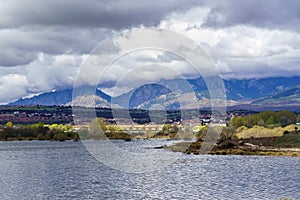 Green landscape with blue lake and houses on the shore, dramatic sky with storm clouds. Guadalix Madrid