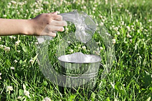 On a green lawn grass with flowers of clover there is a bucket with plastic garbage and a hand collects plastic bottles into it
