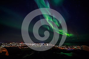 Green lights of Aurora Borealis with shining stars over the fjord and highlighted city, Nuuk, Greenland