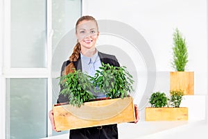 Green living. Attractive young businesswoman standing in modern loft office holding a box with plants. Environmental