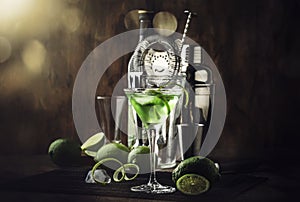 Green martini alcoholic cocktail in glass with dry gin, vermouth, liquor, lime zest and ice, steel bar tools, dark background