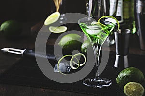 Green martini alcoholic cocktail in glass with dry gin, vermouth, liquor, lime zest and ice, steel bar tools, dark background