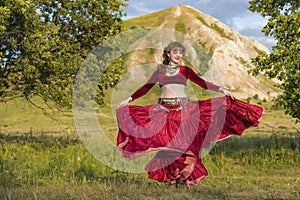 On green meadow against of mountain and summer sunset, young girl in ball gown is dancing incendiary dance.