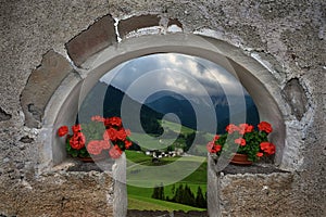 Green meadows, village and mountains through old wall  arch with red flowers.