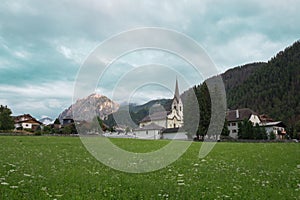 Green Meadows, Wooden Houses and Church in the Village of San Vigilio di Marebbe and Italian Alps Mountains in the Background on a