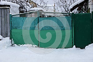 Green metal closed gate and part of the fence on the street in white snow