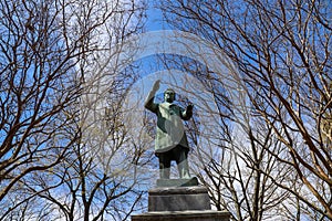 A green metal statue of a man in a overcoat surrounded by bare winter trees and lush green trees with blue sky