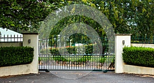 Green metal wrought iron driveway property entrance gates set in concrete  fence, garden shrubs, trees, lights