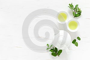 Green mint tea with fresh leaves in cups and teapot overhead on white wooden table, healthy warming drink, antioxidant beverage