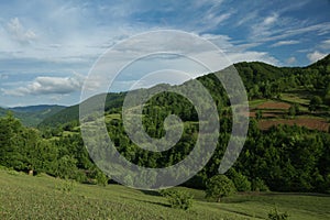 Green mountainsides with trees and a vegetable garden under a blue cloudy sky.