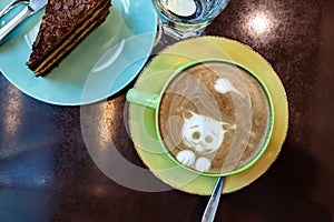 green mug with cappuccino coffee, top view. A cat and a heart are drawn on the foam. On a brown table is a plate with a piece of