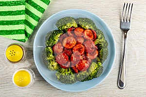 Green napkin, salt and pepper, blue plate with boiled broccoli and slices of sausages, fork on wooden plate