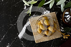 Green olives in a glass bowl and a golden spoon, extra virgin olive oil, and fresh branches on a dark background with copy space