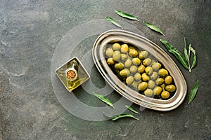 Green olives in olive oil, metal plate, dark rustic background, decorated with leaves. Top view, flat lay, copy space
