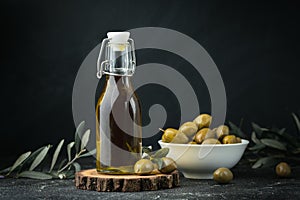 Green olives in a white bowl next to a bottle with olive oil and leaves on a black background. Bottle of cold pressed oil.