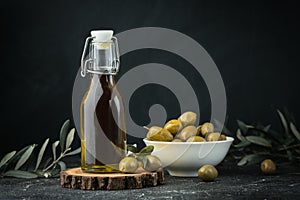Green olives in a white bowl next to a bottle with olive oil and leaves on a black background. Bottle of cold pressed oil.