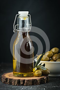 Green olives in a white bowl next to a bottle with olive oil and leaves on a black background. Bottle of cold pressed oil.