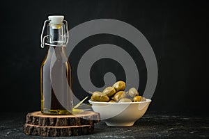 Green olives in a white bowl next to a bottle with olive oil and leaves on a black background. Bottle of cold pressed oil.