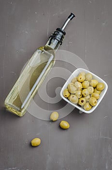 Green olives in a white ceramic bowl and glass bottle of olive oil on a dark background. Top view