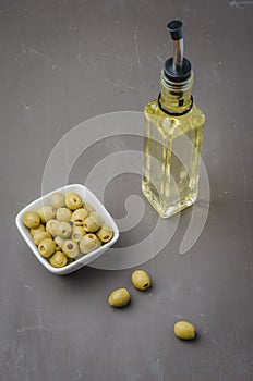 Green olives in a white ceramic bowl and glass bottle of olive oil on a dark background. Top view