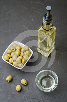 Green olives in a white ceramic bowl and glass bottle of olive oil on a dark background. Top view