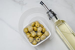 Green olives in a white ceramic bowl and glass bottle of olive oil on a light concrete background. Top view