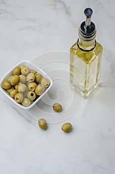Green olives in a white ceramic bowl and glass bottle of olive oil on a light concrete background. Top view