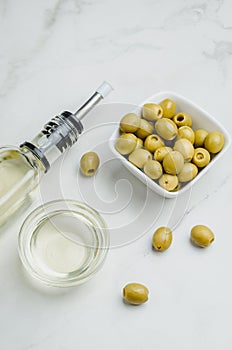 Green olives in a white ceramic bowl and glass bottle of olive oil on a white background. Top view