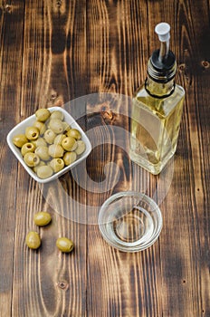 Green olives in a white ceramic bowl and glass bottle of olive oil on a wooden background. Top view