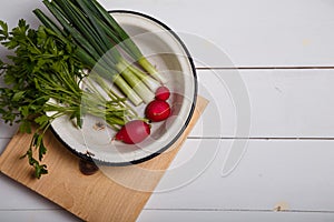 Green onions, parsley, radish in white metallic bowl on kitchen cutting board on wooden table. Fresh vegetables and greenery