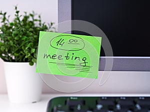 A green paper notes with the reminder 14-00 Meeting on it sticked on to a monitor at an office workplace