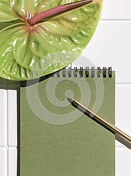 Green paper spiral notepad with anthurium or flamingo flower, pen on the white ceramic tile table