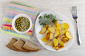 Green peas in bowl, bread, plate with slices of fried potatoes and dill, fork on table. Top view