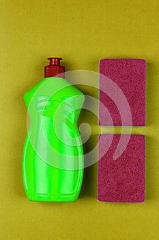 Green plastic bottle of dishwashing liquid and red sponge on greenish-yellow background close-up top view,
