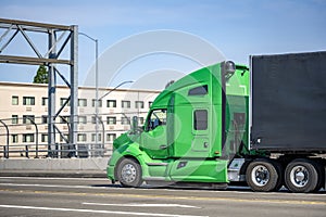 Green professional big rig bonnet semi truck transporting cargo in covered black semi trailer running on the city street bridge