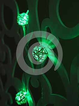 Green Rattan ball garland on patterned wall. Christmas and New Year led lights on dark background. Blurred glowing light bulb