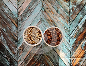 Green raw beans and roasted coffee beans in two white cups on turquoise wooden background top view
