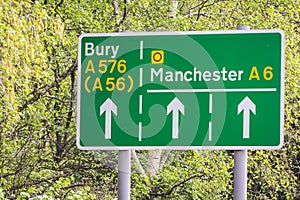 green and white street sign amongst a background of trees with directional guides on the A6 towards manchester or bury