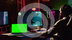 Green screen laptop next to man using gaming keyboard to play spaceship flying singleplayer game